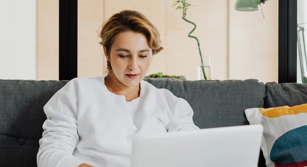Stock photograph: a woman sitting on a grey sofa with a laptop open on her knees, reading in a room with tall windows behind her